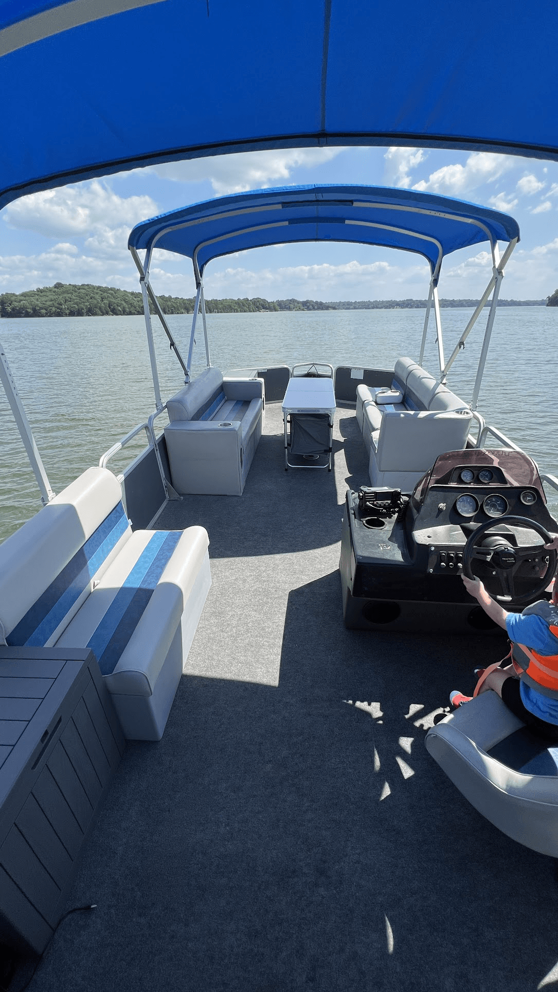 Pontoon boat interior featuring blue canopies and grey seating on a calm, sunny lake.