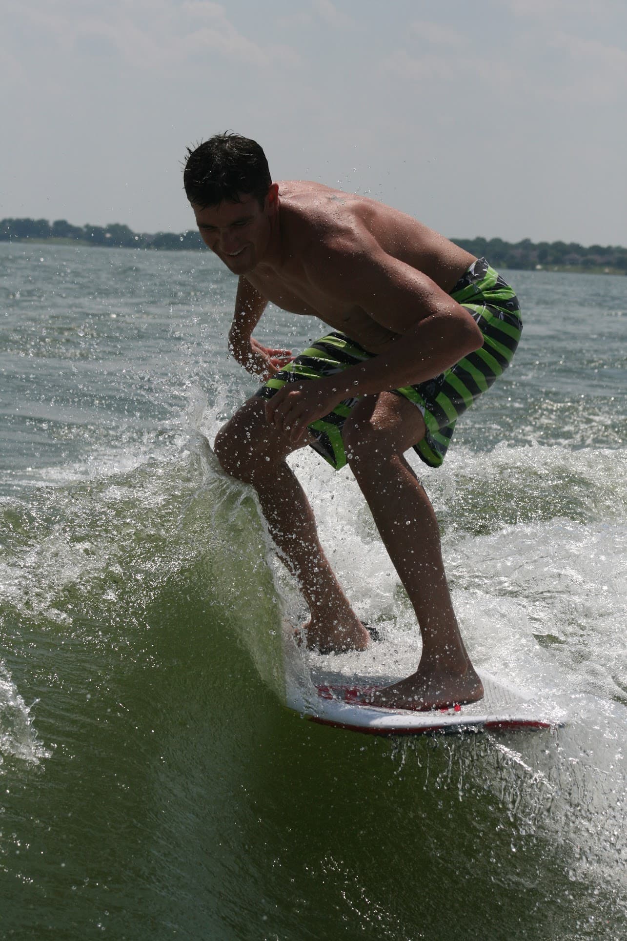 Shirtless man in green and black striped shorts wakesurfing on a splashing lake wave.