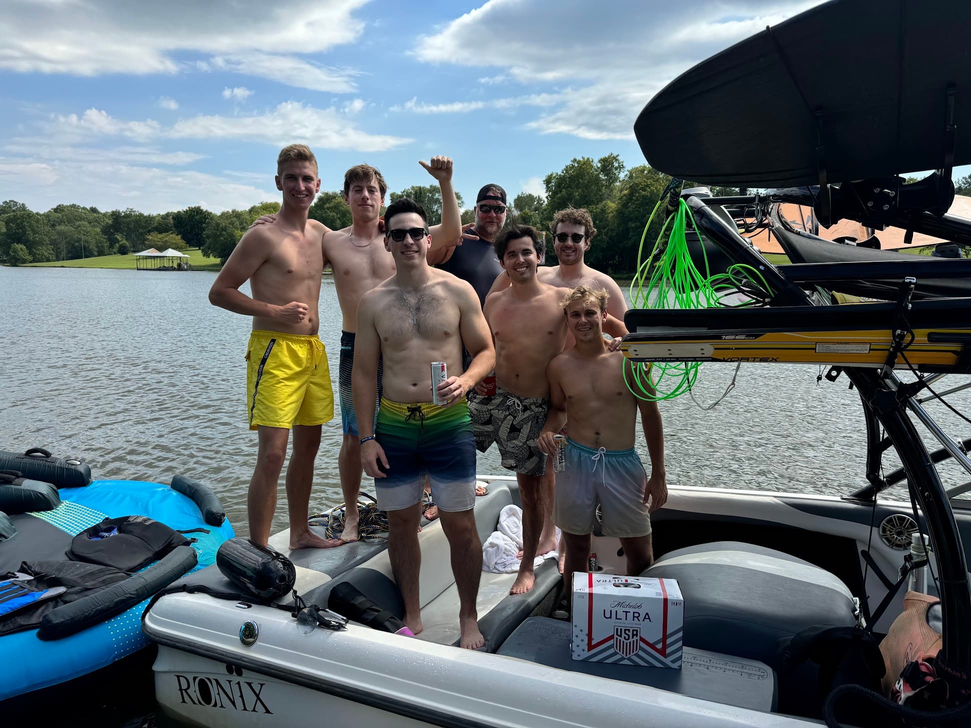 Seven men in swim trunks posing on a motorboat on a lake during the summer.